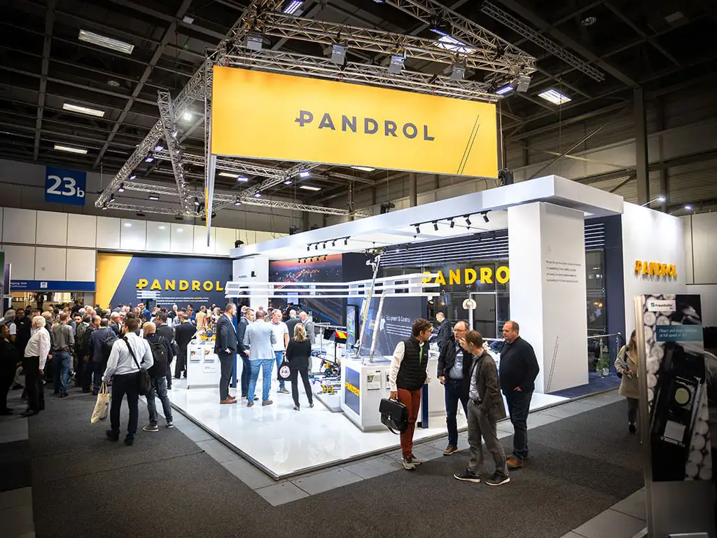 Pandrol's exhibition stand, featuring a large hanging sign in yellow with 'Pandrol' on each side. Many visitors crowd around different areas of the stand