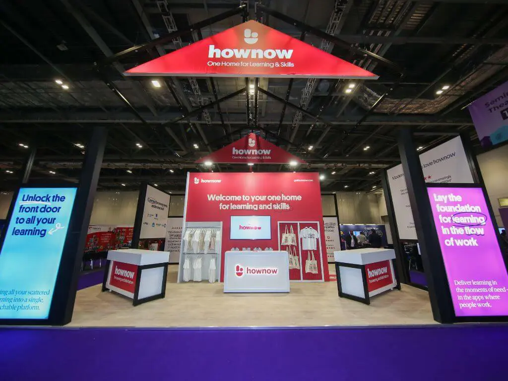 A bespoke exhibition stand build for HowNow at the Learning Technologies event, featuring an apex roof and red and black branding throughout the build elements